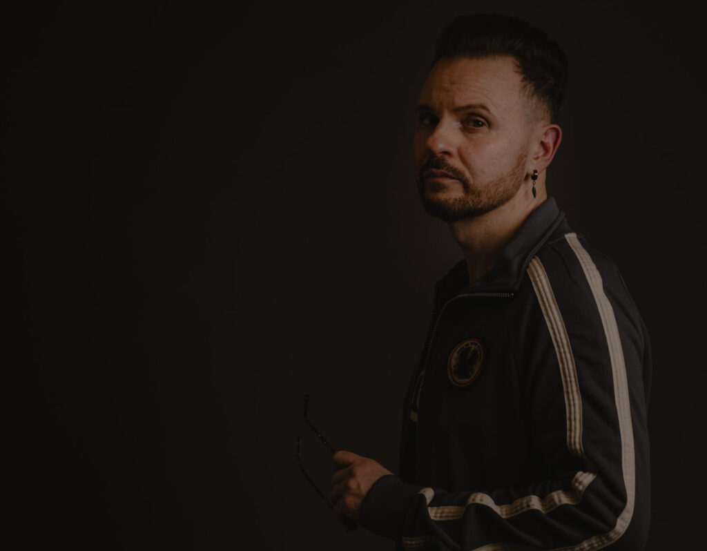 Man in a black tracksuit with white stripes, holding glasses, against a dark studio backdrop.