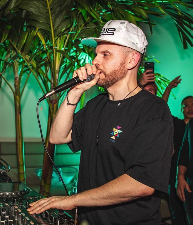 DJ in a white camo cap performing with a microphone, hands on a mixer, green club lighting in the background, audience behind him.