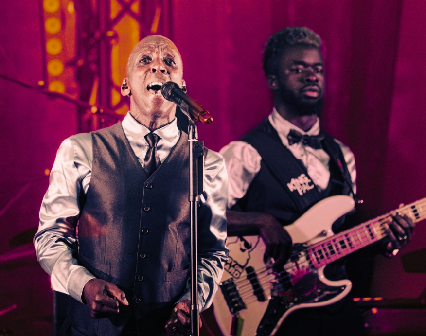 Lead male singer in a gray vest and tie sings into a microphone on a pink-lit stage, with a bassist in the background playing a white bass.