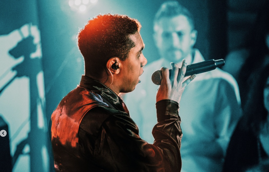 Male singer on stage, microphone in hand, wearing a dark jacket as blue stage lights illuminate the scene.