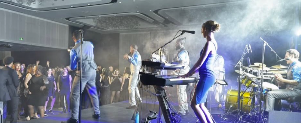 Live band on stage performing for an engaged crowd; keyboardist in blue dress front and center, drummer and singers in background.