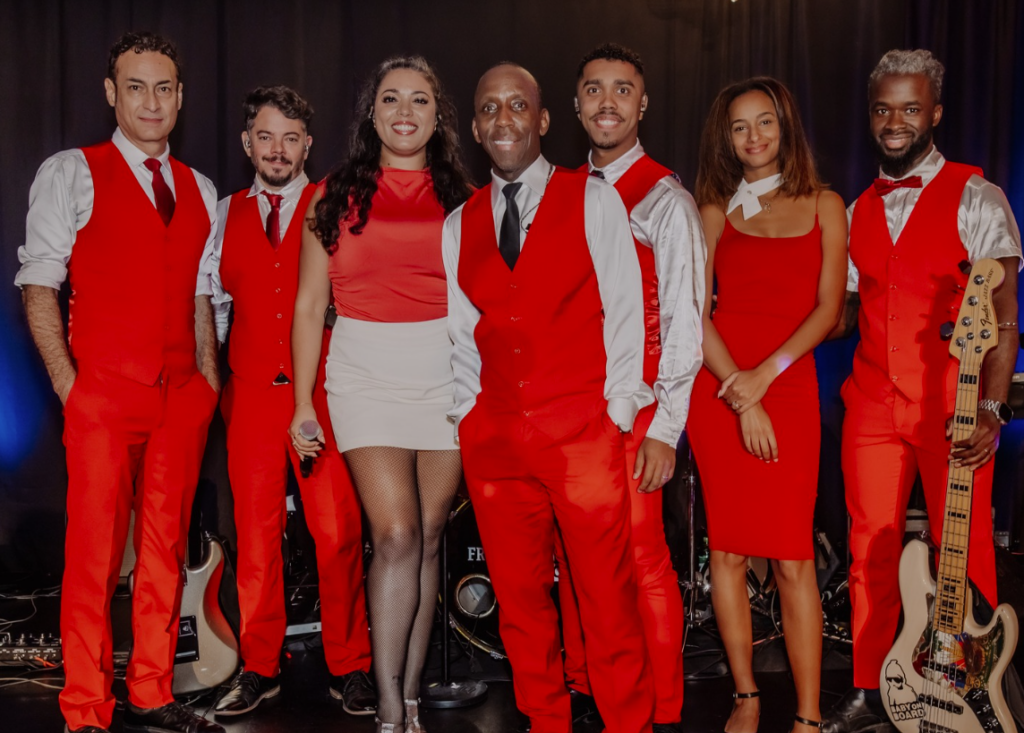 Seven performers pose on a stage in bright red outfits; instruments and a drum set are visible in the background, smiling at the camera.