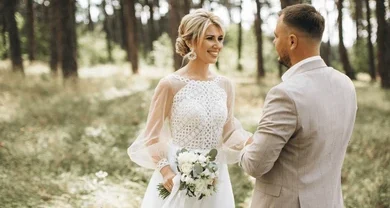 Bride in a white wedding dress holds a bouquet as she faces her groom in a light suit in a sunlit forest setting.