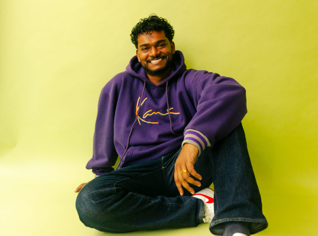 Man with curly hair in a purple hoodie sits against a bright yellow background, smiling at the camera.