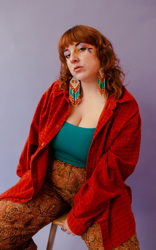 Woman with red hair in a teal top and red jacket, wearing large colorful beaded earrings against a lavender background.
