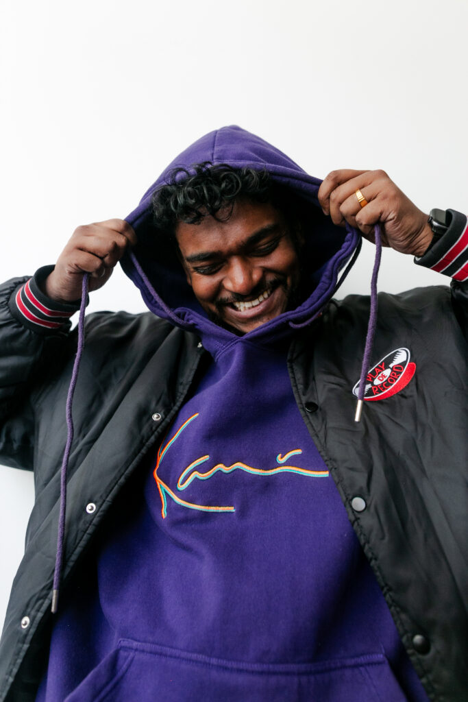 Man smiles while pulling up a purple hoodie over his head, wearing a black jacket with striped cuffs and a logo patch on the chest