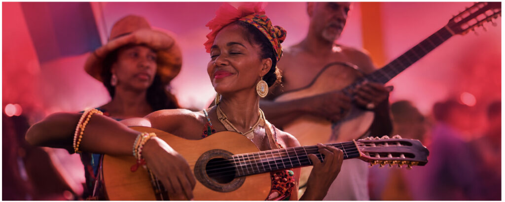 Smiling woman strums a guitar at a colorful outdoor festival, with other musicians in the pink-lit background, enjoying the moment.