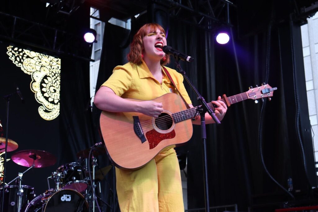 Female singer in a yellow outfit performing on stage, guitar in hand, singing into a microphone.